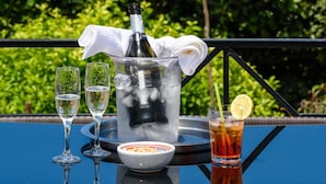 Een tafel met een champagnefles, twee glazen, een karaf, een schaal en een theeglas, voor een achtergrond van weelderige groene begroeiing en een bergketen in de verte.
