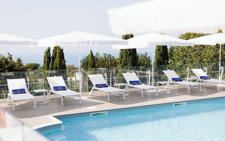 A poolside area with white lounge chairs and umbrellas, surrounded by a glass barrier and overlooking a landscape with trees and a clear sky.