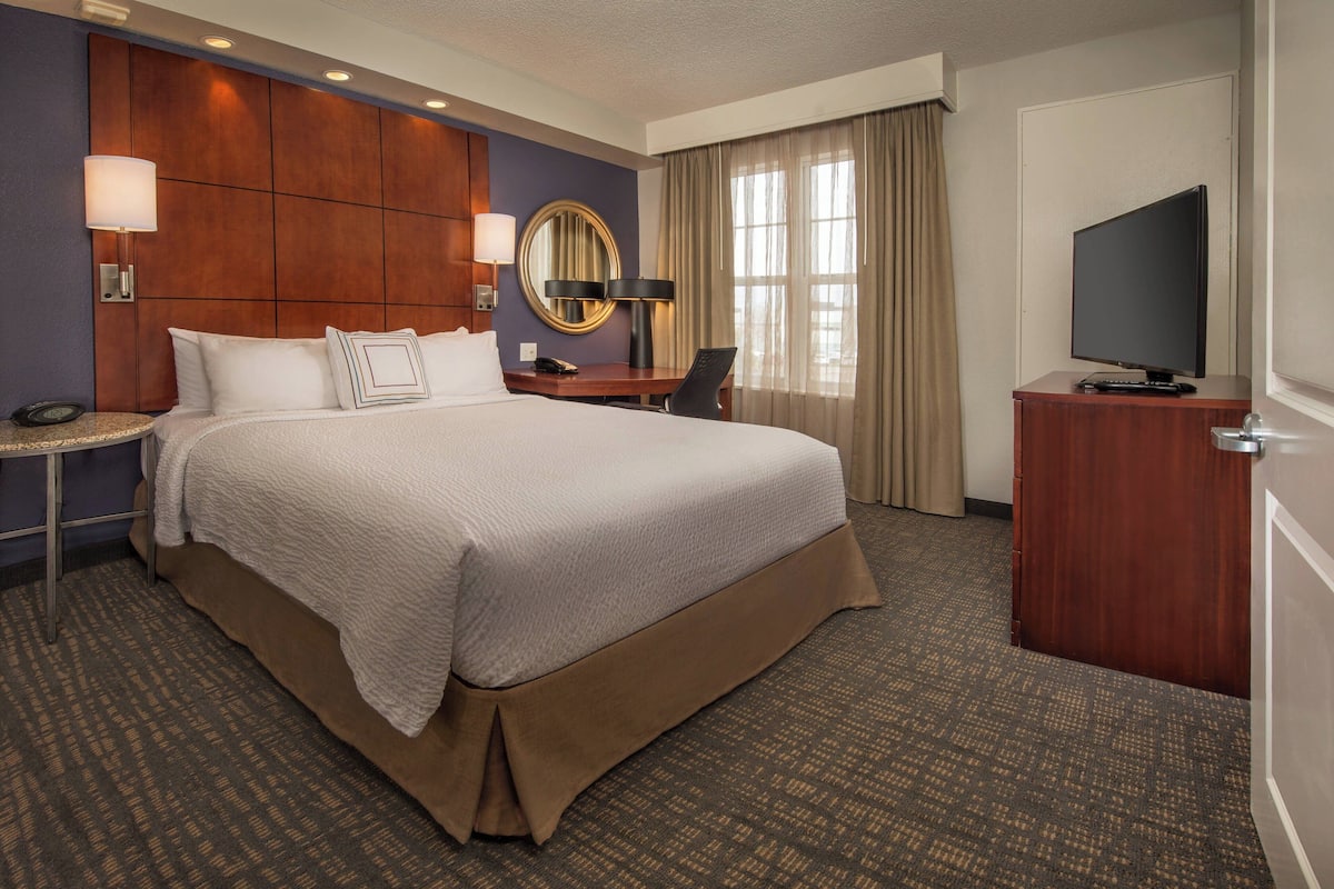 A hotel room with a large bed, a desk with a chair, a television, a mirror, and a lamp.