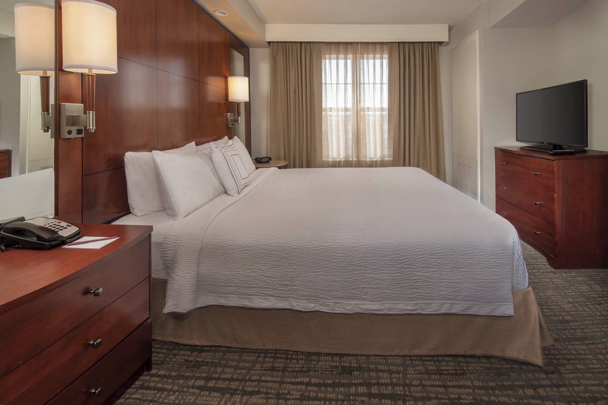 A hotel room with a large bed, wooden headboard, bedside lamps, a flat-screen TV, and a telephone on a wooden dresser.