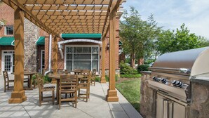 Un patio al aire libre con mesas y sillas de madera, una parrilla y un área de estar cubierta con un muro de piedra de fondo.