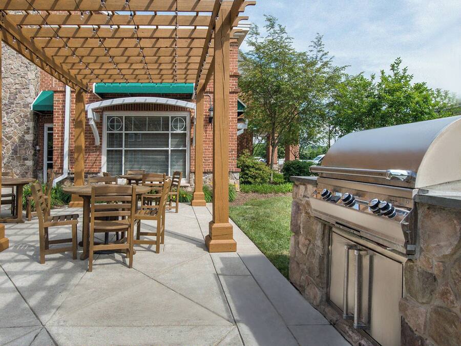 An outdoor patio with wooden tables and chairs, a barbecue grill, and a covered seating area with a stone wall backdrop.