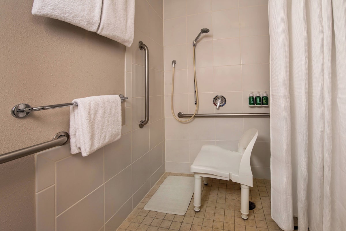 A bathroom with a shower, grab bars, a white sink, and a white chair.