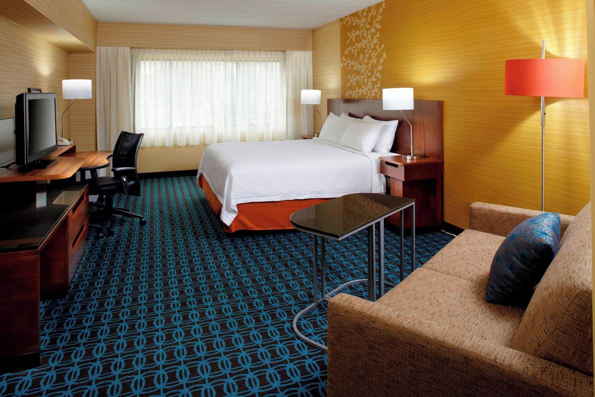 Ein Hotelzimmer mit einem Bett, einem Schreibtisch mit Computer, einem Sessel und einem Sofa sowie einem Fernseher.