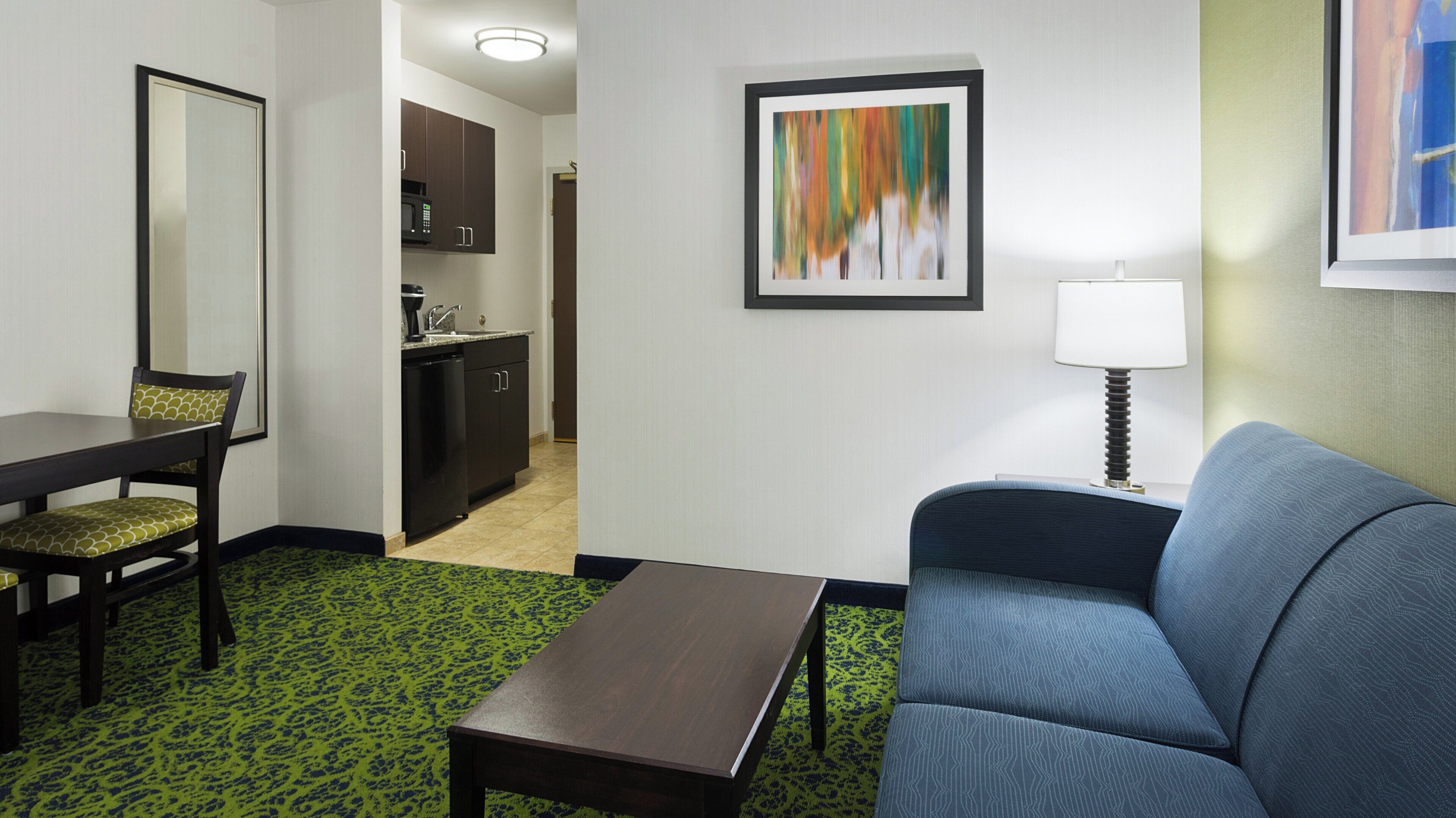 A hotel room with a dining table, a sofa, a coffee table, a kitchen area, and a framed abstract painting on the wall.