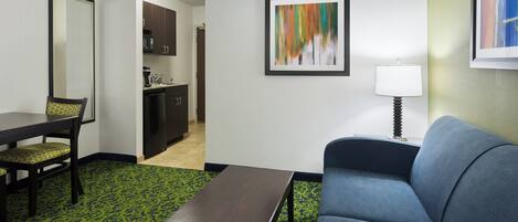 A hotel room with a dining table, a sofa, a coffee table, a kitchen area, and a framed abstract painting on the wall.