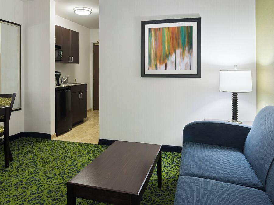 Habitación de hotel con una mesa de comedor, un sofá, una mesa baja, una zona de cocina y un cuadro abstracto enmarcado en la pared.