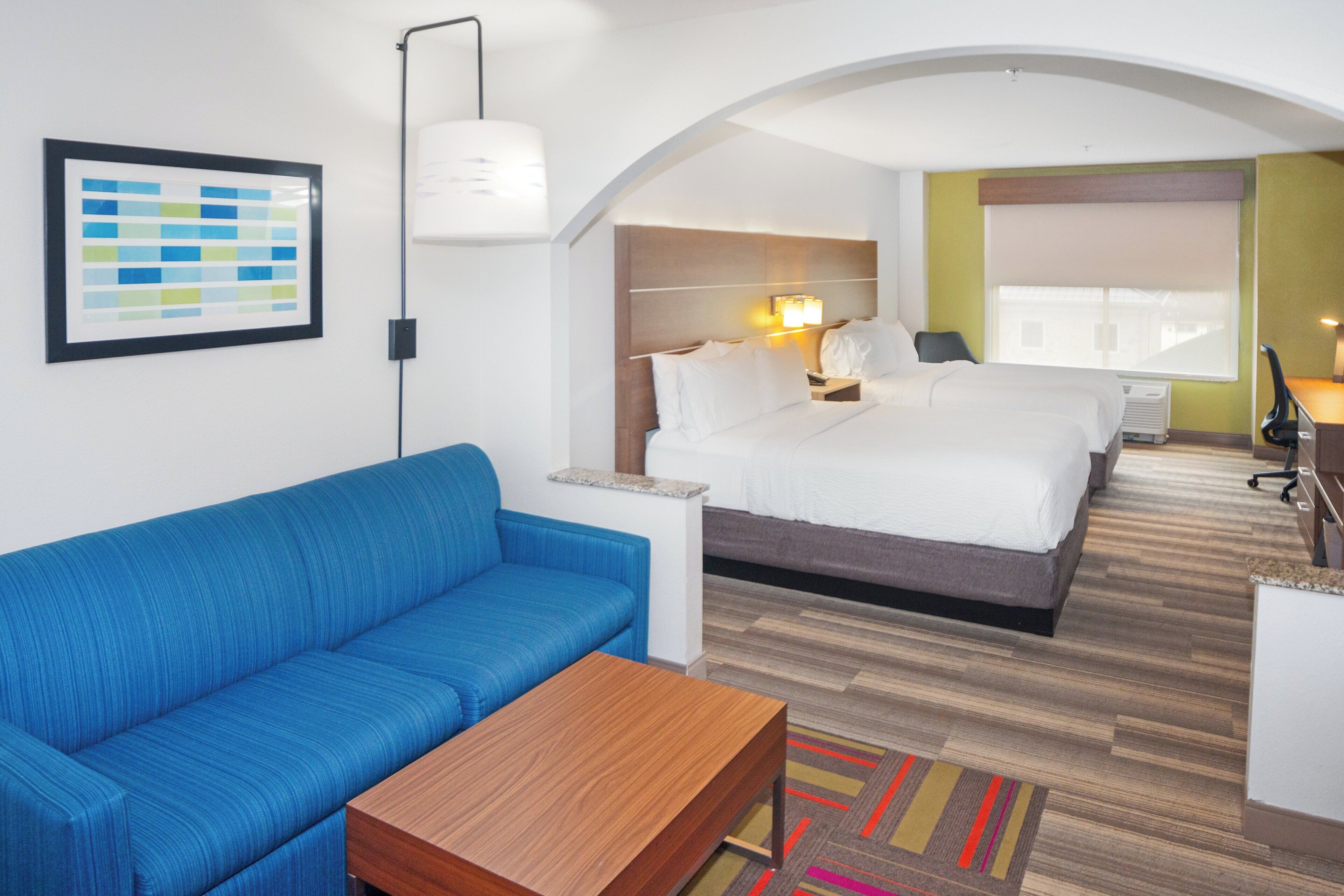 A hotel room with a blue sofa, a wooden coffee table, a bed with white linens, a nightstand, a desk with a chair, and a colorful rug.
