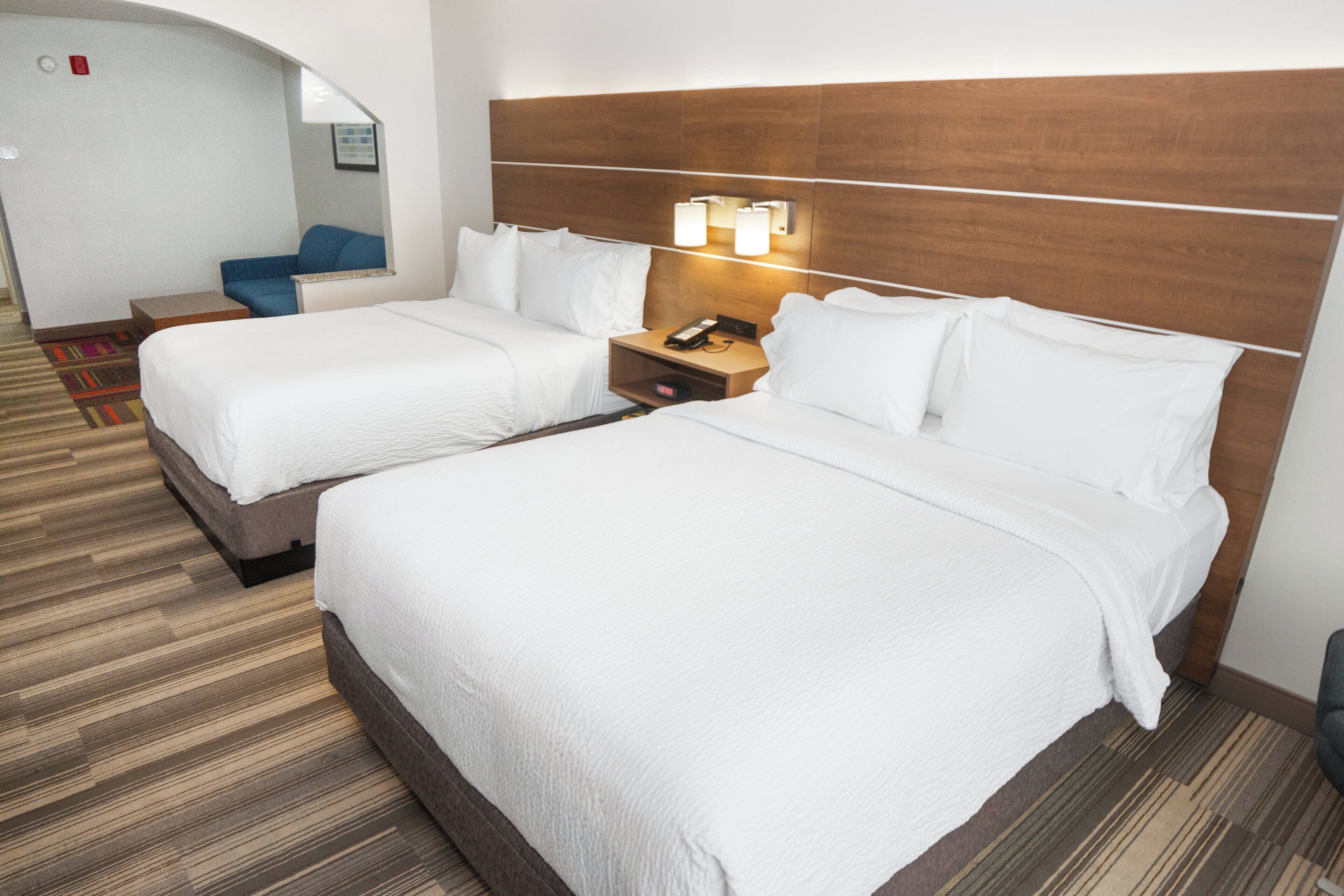 A hotel room with two beds, a nightstand with a phone, and a seating area with a chair and a small table.
