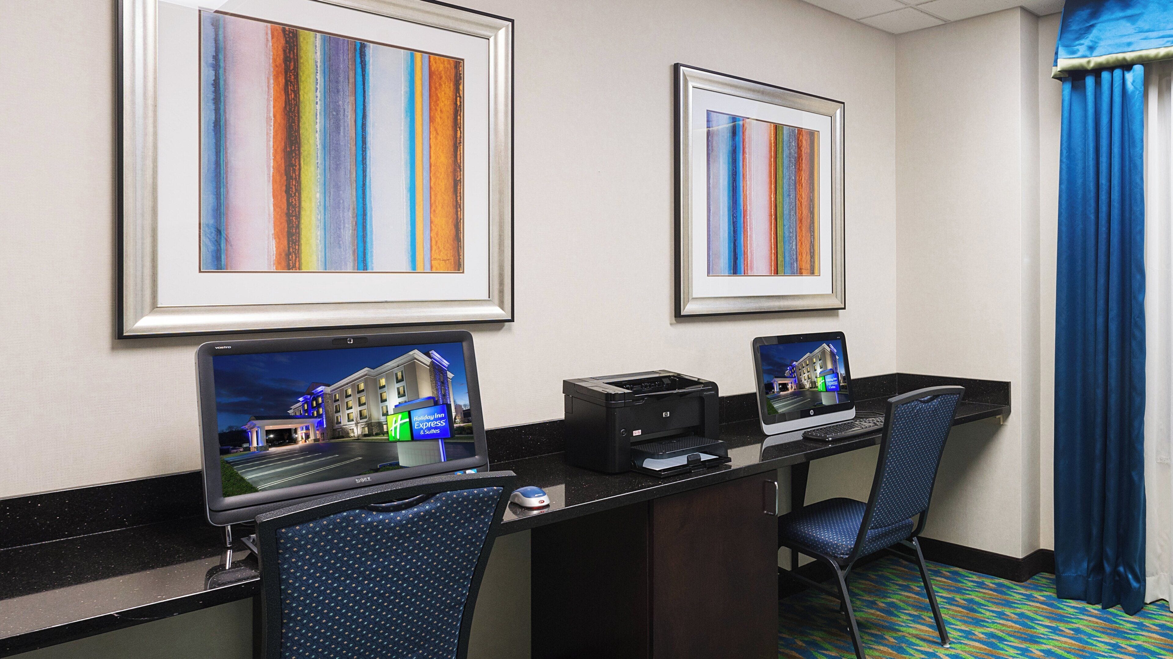 A hotel room with two desks, each with a computer monitor, printer, and keyboard. There are framed pictures on the wall and a blue curtain on the window.