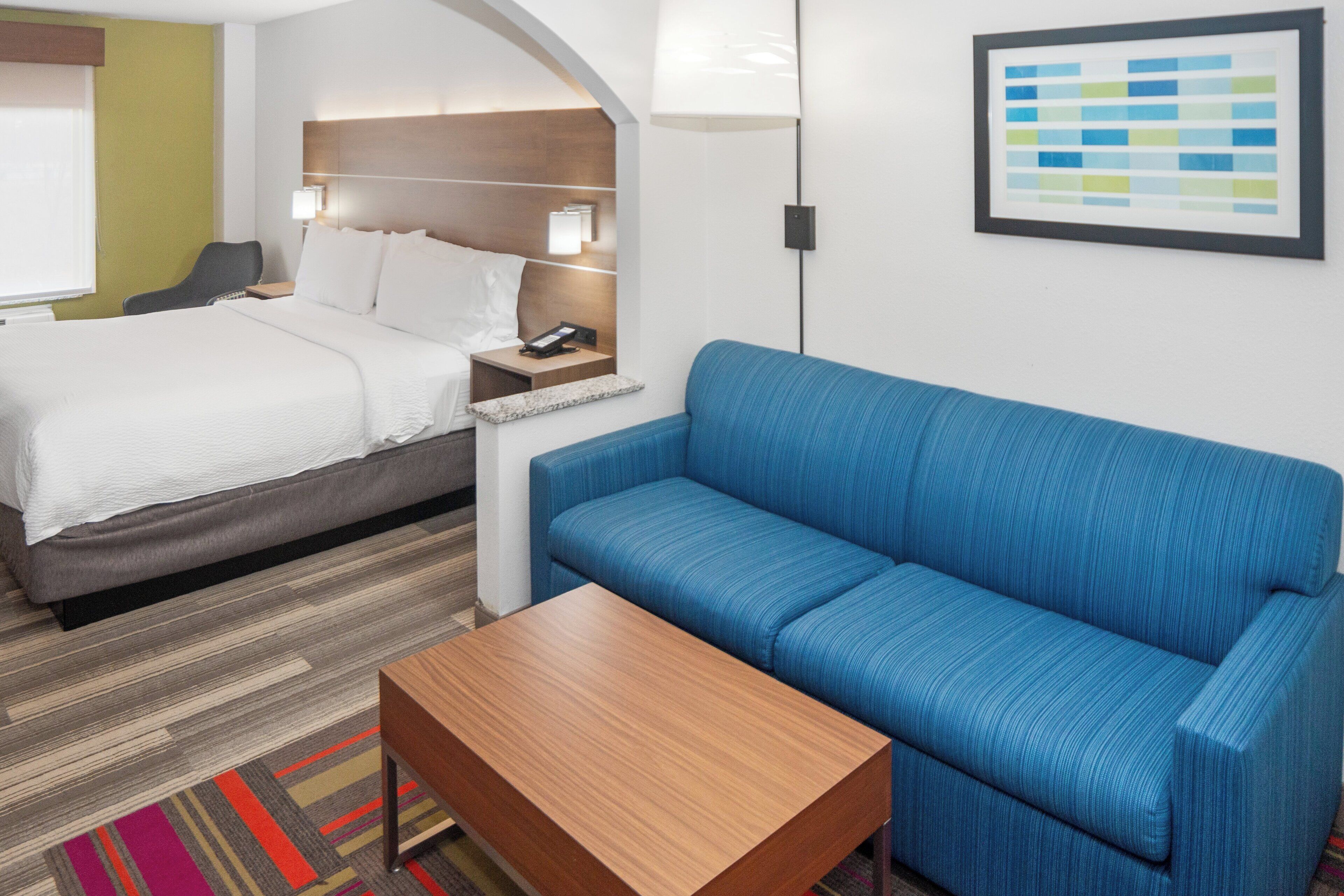 A hotel room with a bed, a blue sofa, a wooden coffee table, and a nightstand with a lamp.