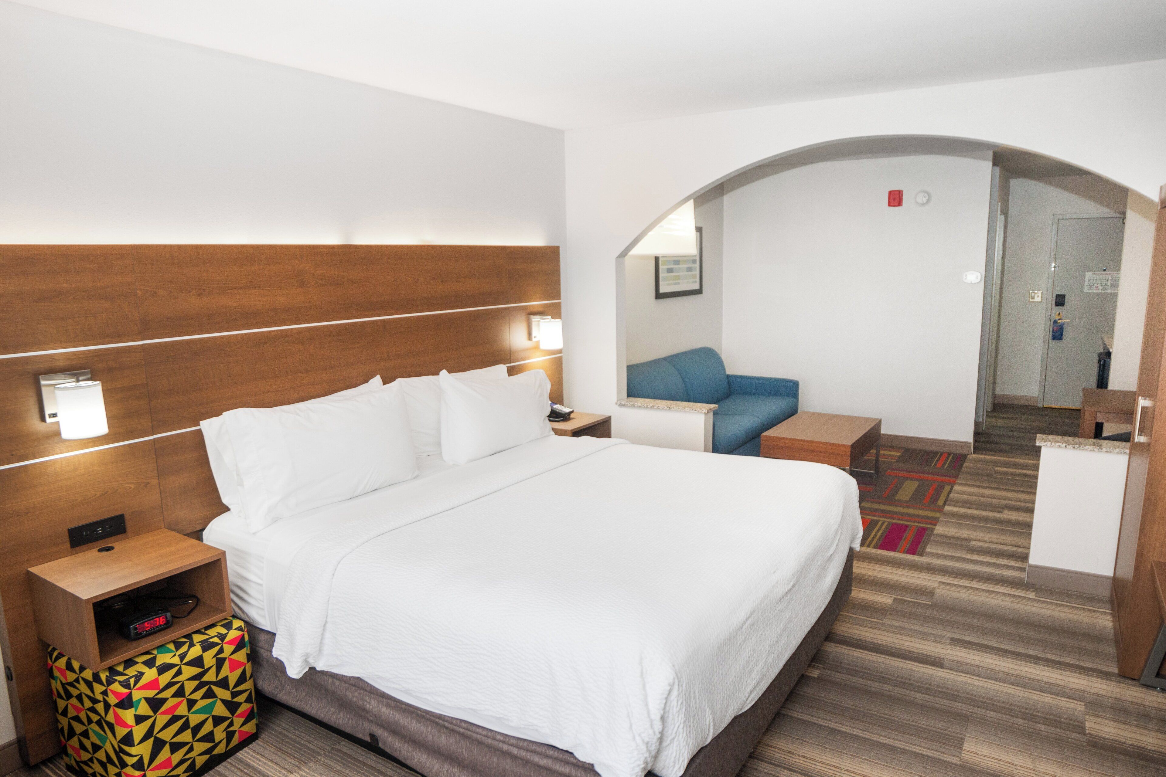 A hotel room with a large bed, bedside tables, a small desk, a colorful bag, and a mirror.