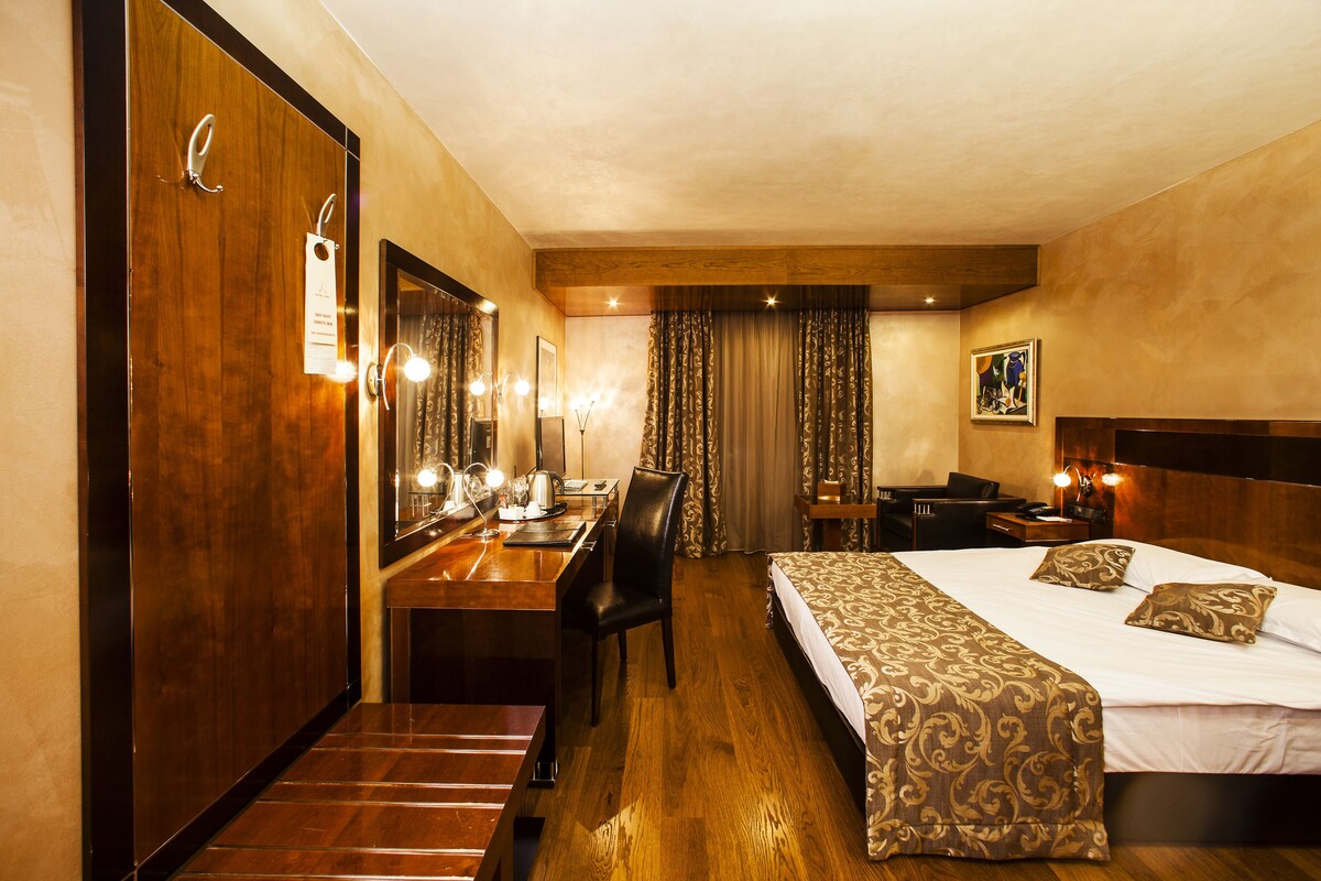 A hotel room with a wooden desk, a large mirror, a bed with patterned bedding, and a headboard.