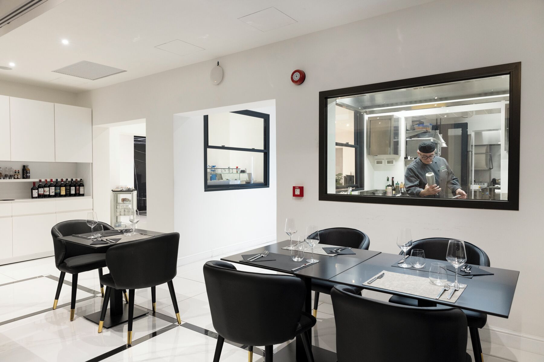 A modern dining area with a large table, black chairs, and wine glasses. A chef is visible in the reflection of a large mirror.