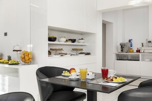 A modern kitchen with white cabinetry, a dining table with chairs, and a breakfast setting with fruit and beverages.