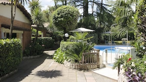 Un área de piscina con un sendero de piedra, rodeada de plantas y árboles, y un edificio con techo de tejas.