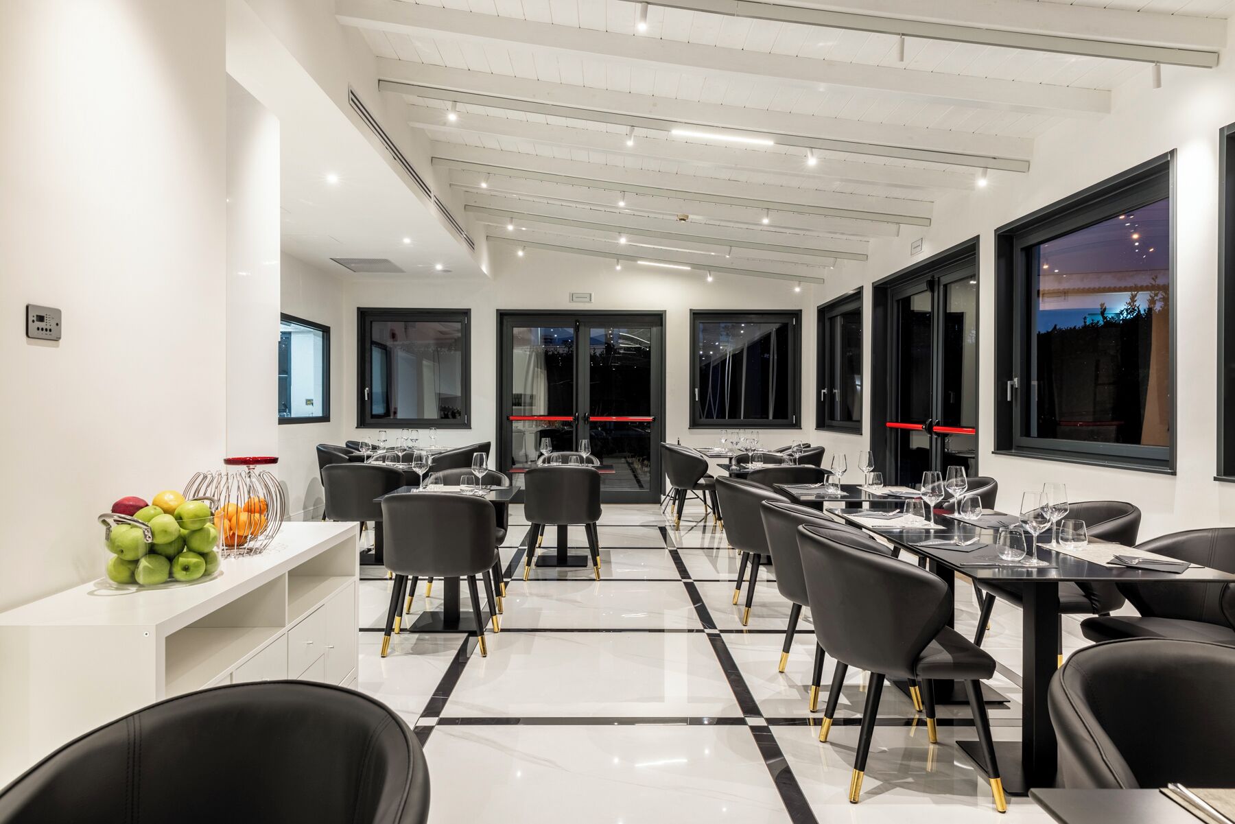 Un comedor moderno con sillas y mesas negras, una exhibición de frutas y grandes ventanales con vistas a la ciudad.