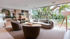 A modern living room with a sofa, armchairs, and a coffee table, a bookshelf with plants, and a large window with a view of greenery. - BLUESEA Club Martha’s (Santanyi)