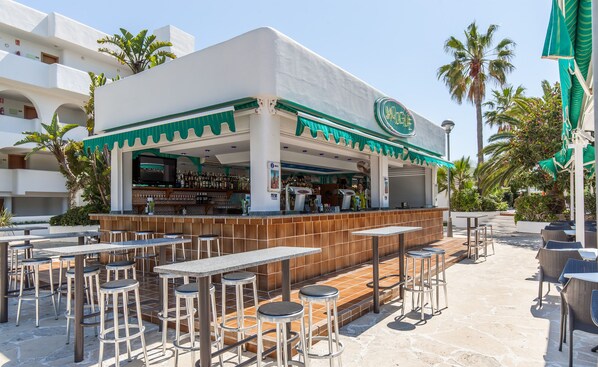 An outdoor bar area with high stools and tables, surrounded by palm trees and a white building with green accents. - BLUESEA Club Martha’s (Santanyi)