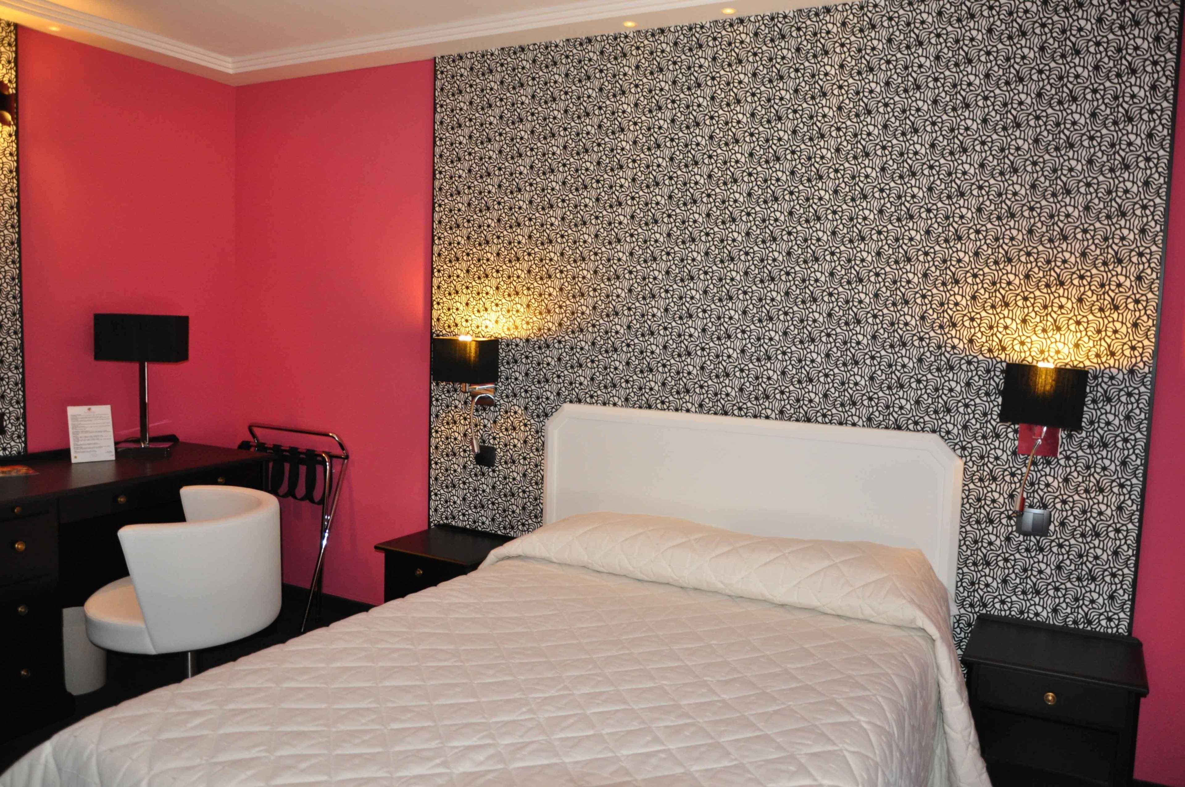 A hotel room with a bed, a desk with a chair, a lamp, and a wall with a patterned wallpaper.