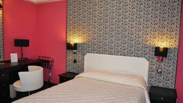 A hotel room with a bed, a desk with a chair, a lamp, and a wall with a patterned wallpaper.