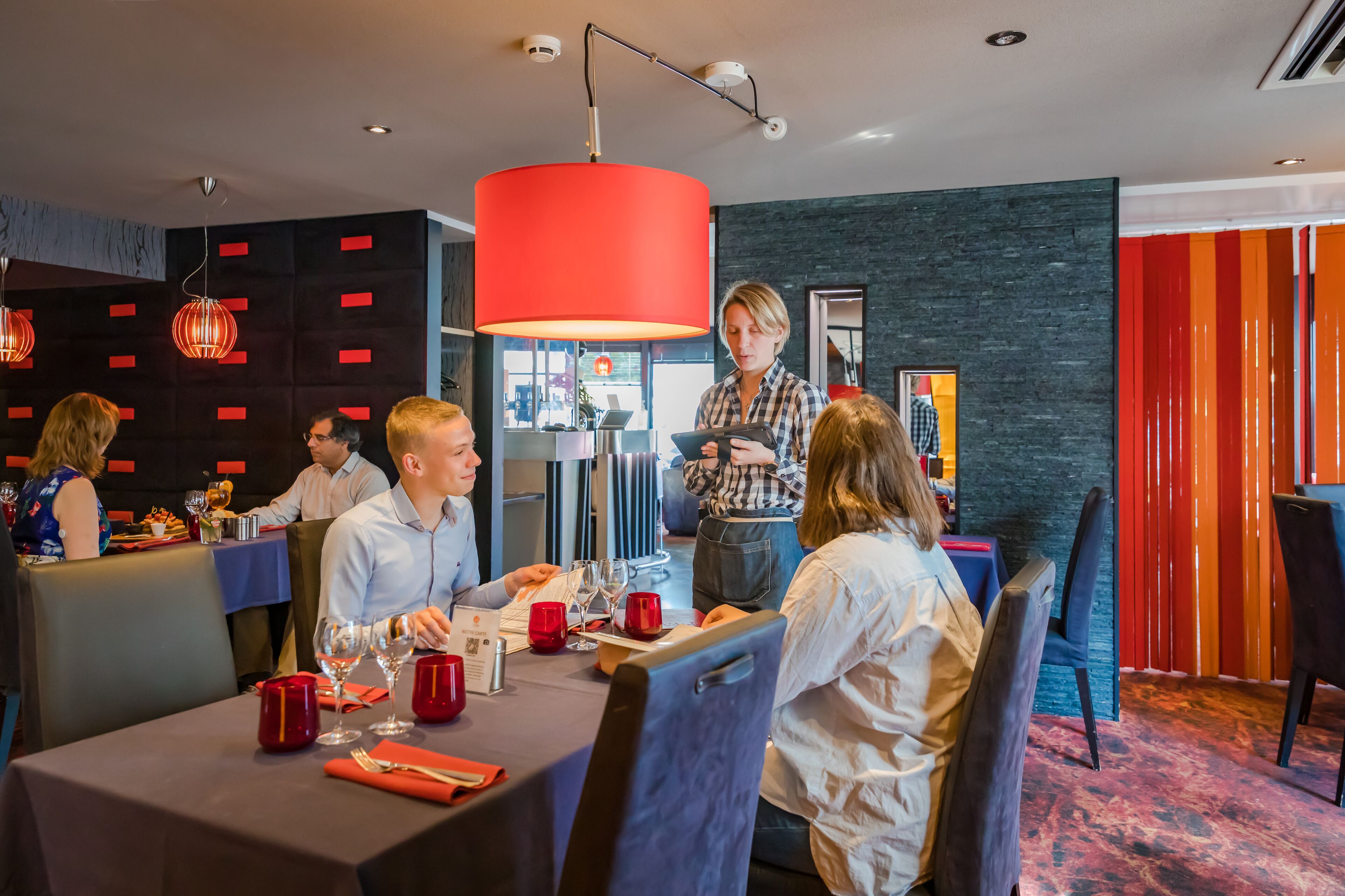 A modern dining area with tables set for a meal, a large red pendant light, and a person standing by the table.
