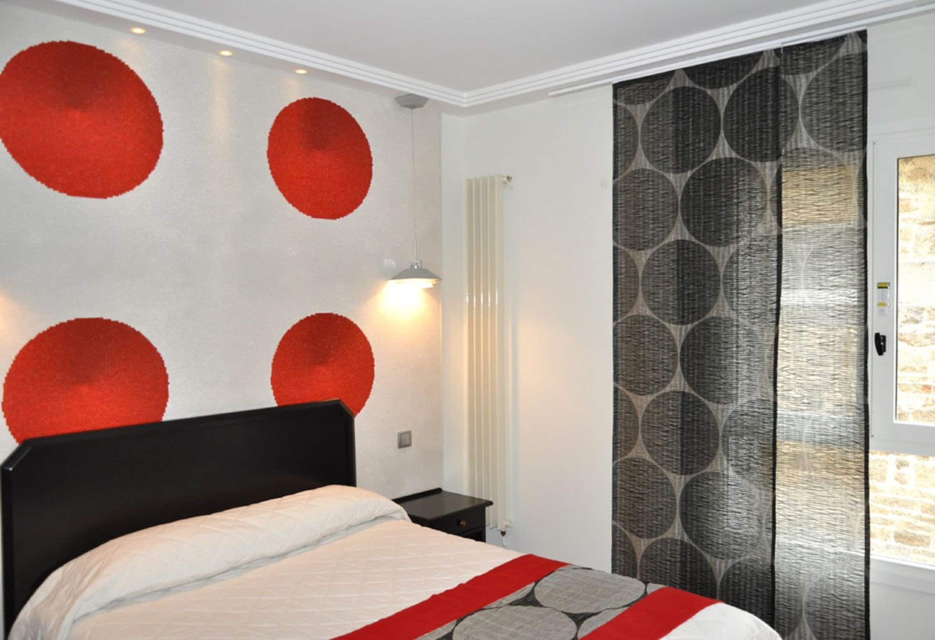 Een slaapkamer met een bed, nachtkastje, een wandlamp en een raam met gordijnen met patroon.