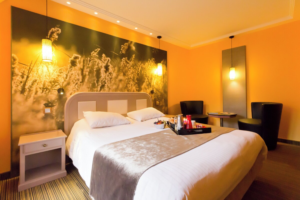 A hotel room with a large bed, bedside tables, a nightstand, a seating area with a chair and table, and a wall-mounted light fixture.
