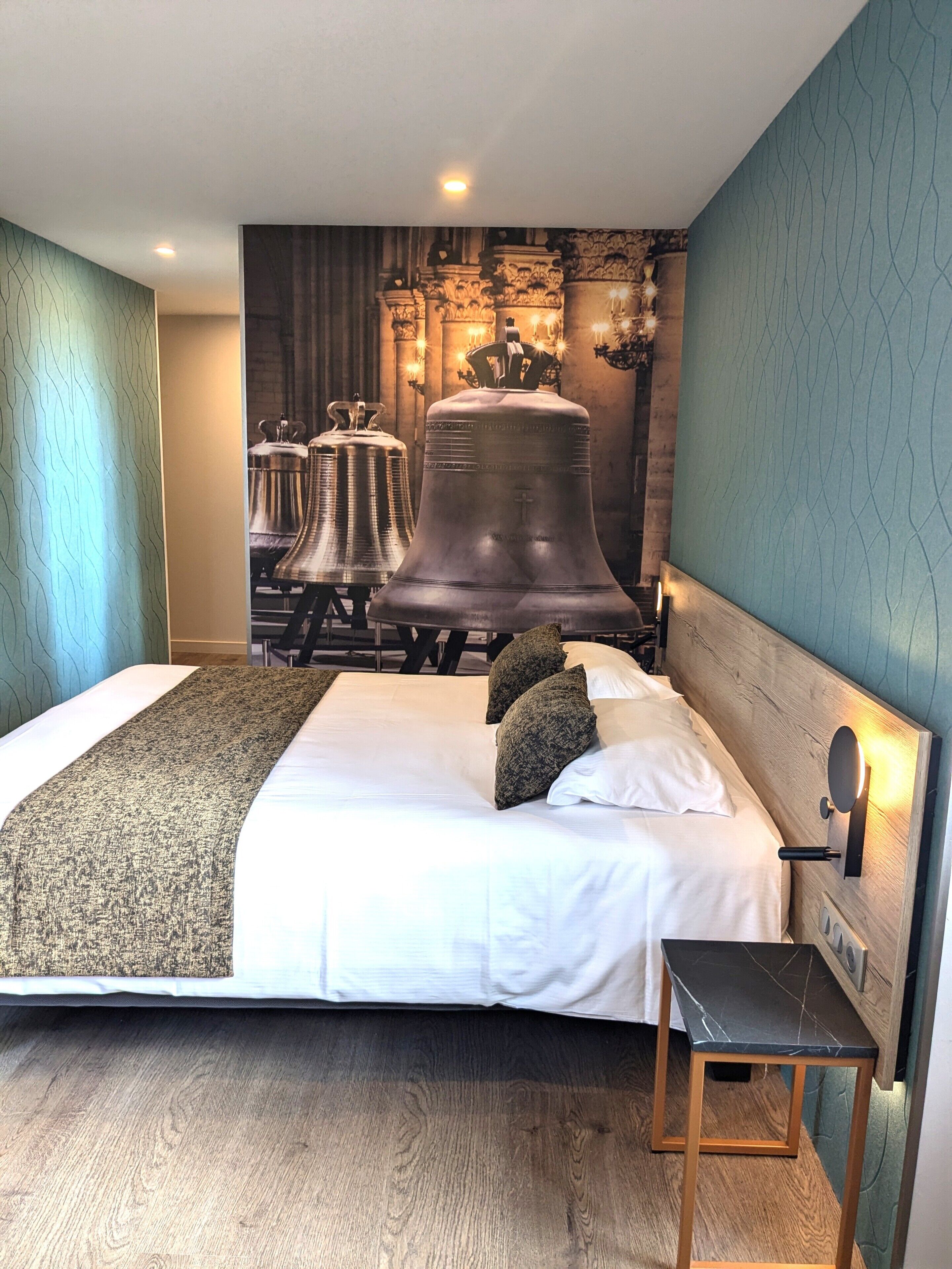 A large bed with a headboard, a bedside table, and a wall with a bell mural.