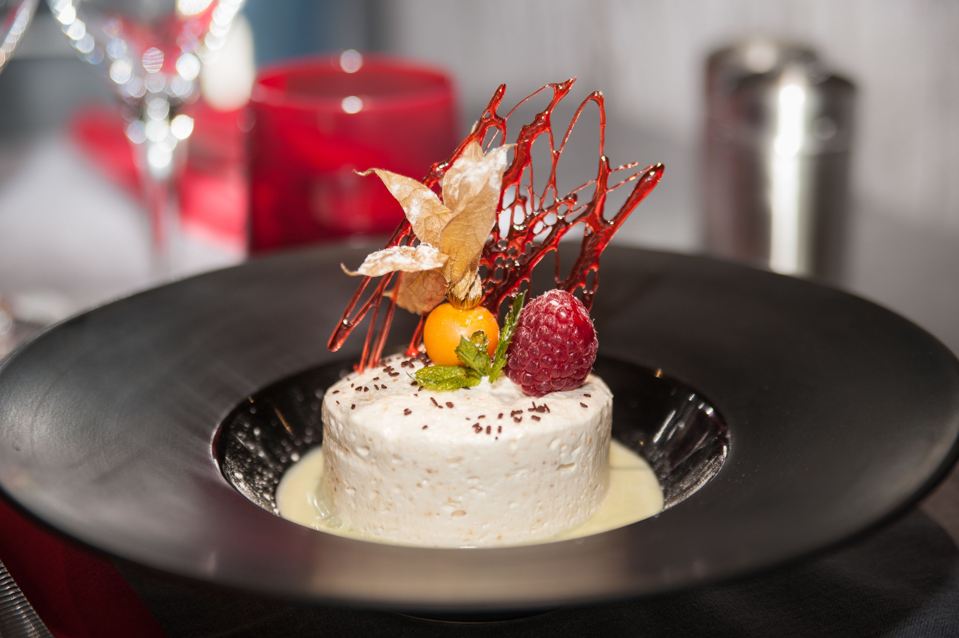 A dessert on a black plate with a white base, garnished with a leaf, a raspberry, and a small orange.