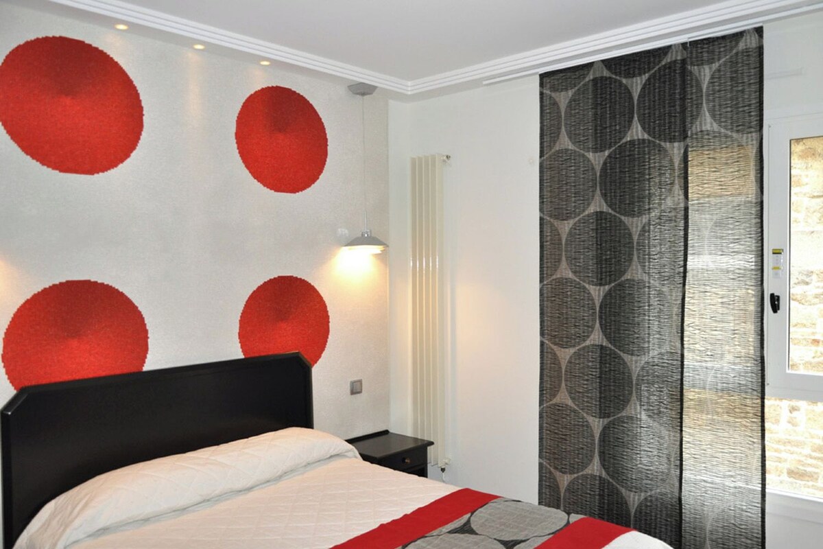 A bedroom with a bed, a nightstand, a wall with circular patterns, and a window with patterned curtains.