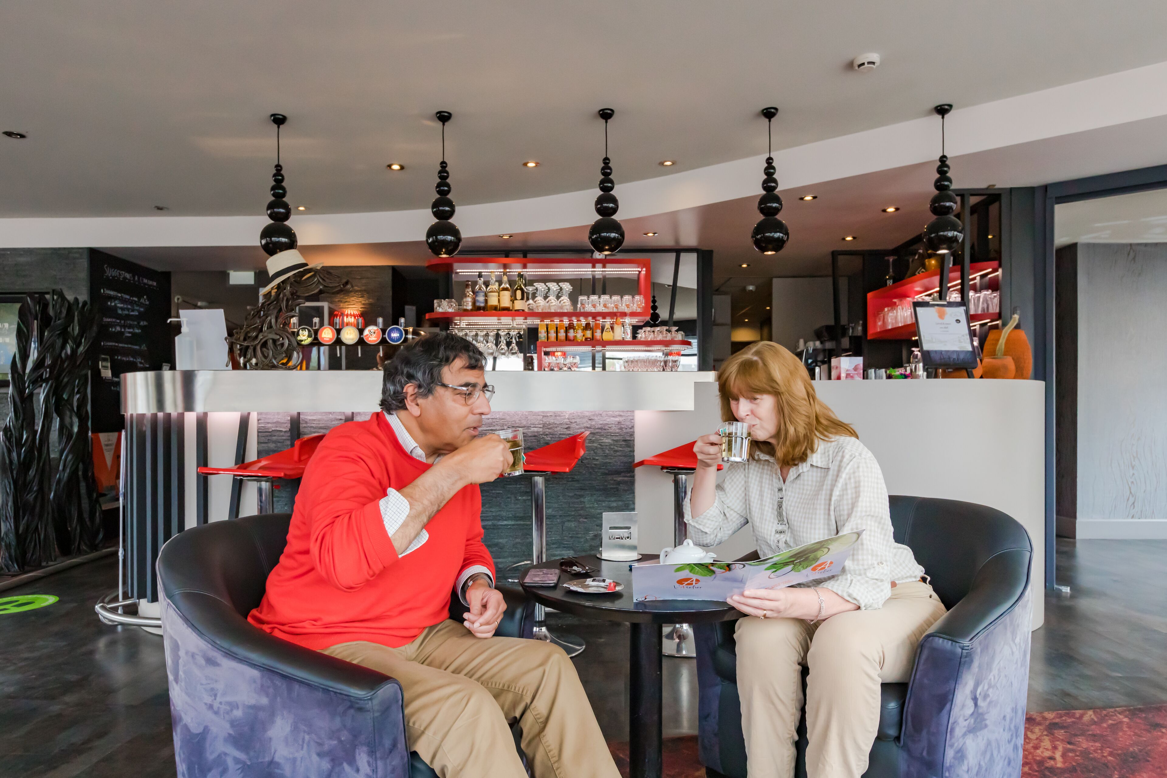 Een moderne bar met een toonbank waarop verschillende flessen en glazen staan. Twee personen zitten aan een tafel met een menukaart en een theepot.