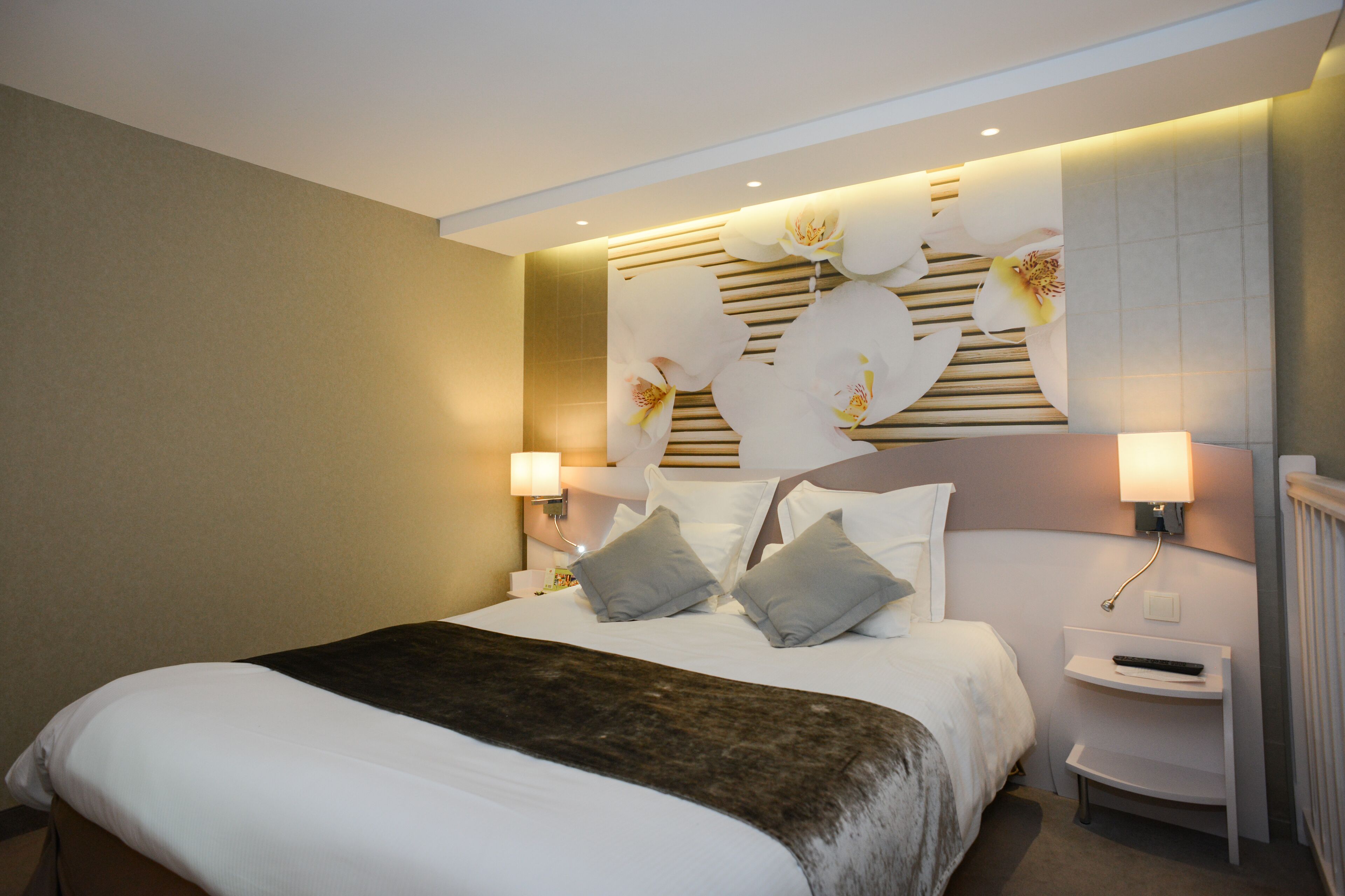Une chambre d’hôtel avec un grand lit, deux tables de chevet, une tête de lit ornée de motifs floraux et une lampe fixée au mur.