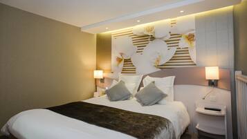 Une chambre d’hôtel avec un grand lit, deux tables de chevet, une tête de lit ornée de motifs floraux et une lampe fixée au mur.
