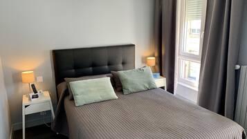 A neatly made bed with a dark headboard, two pillows, and a bedside table with a lamp and a remote control.