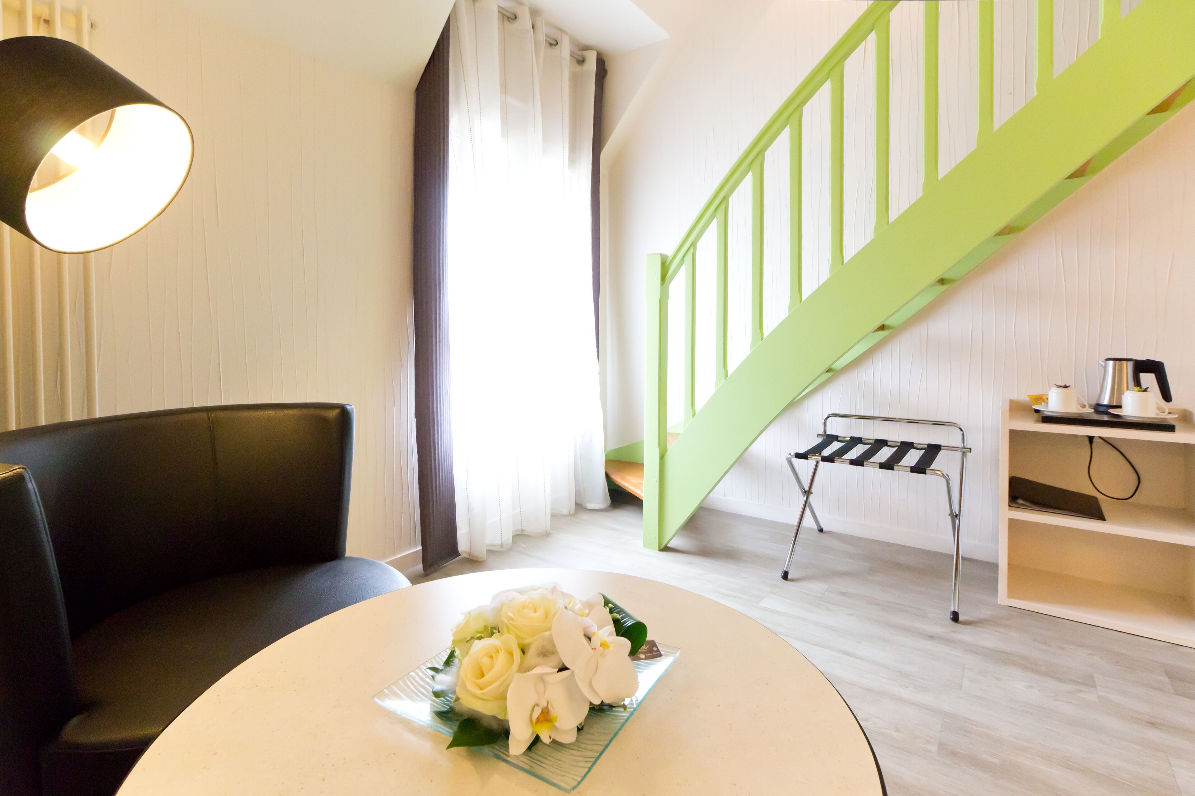 Un salon moderne avec un escalier vert, un canapé en cuir noir, une table ronde blanche agrémentée d’un arrangement floral et une machine à café posée sur une étagère.