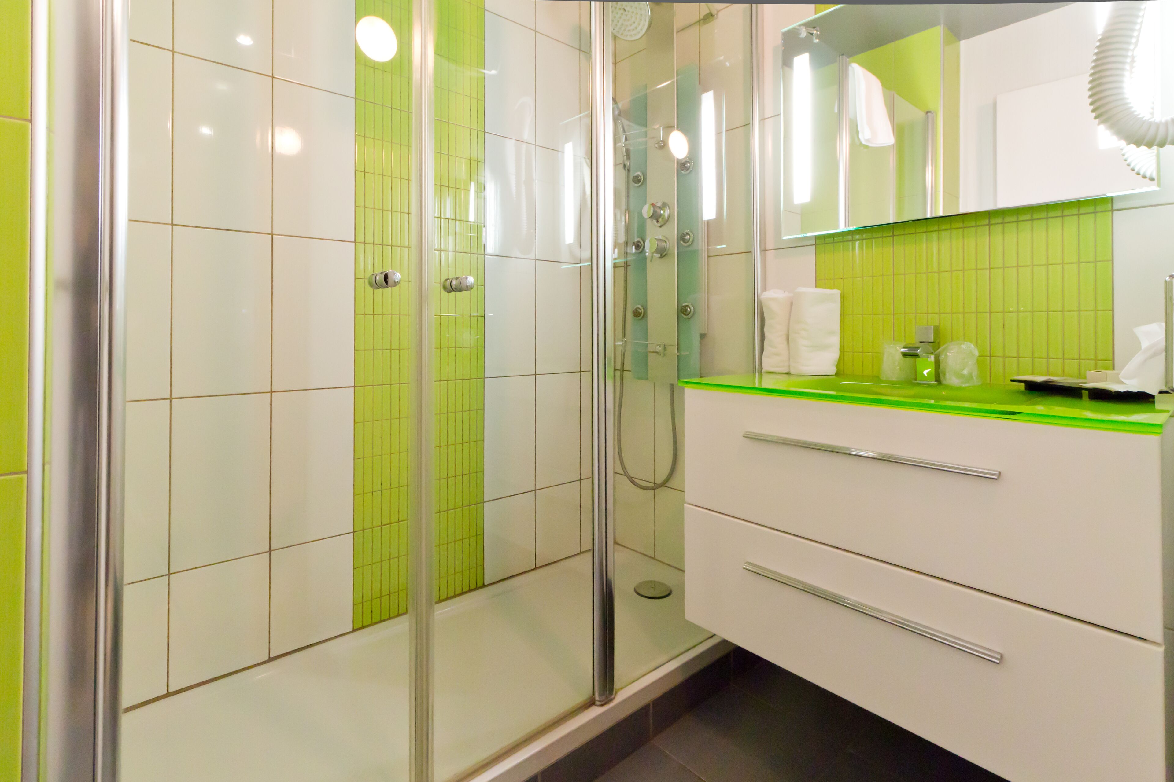 Une salle de bain moderne avec une douche à l’italienne en verre, un mur recouvert de carreaux verts et un plan de toilette blanc surmonté d’un miroir.