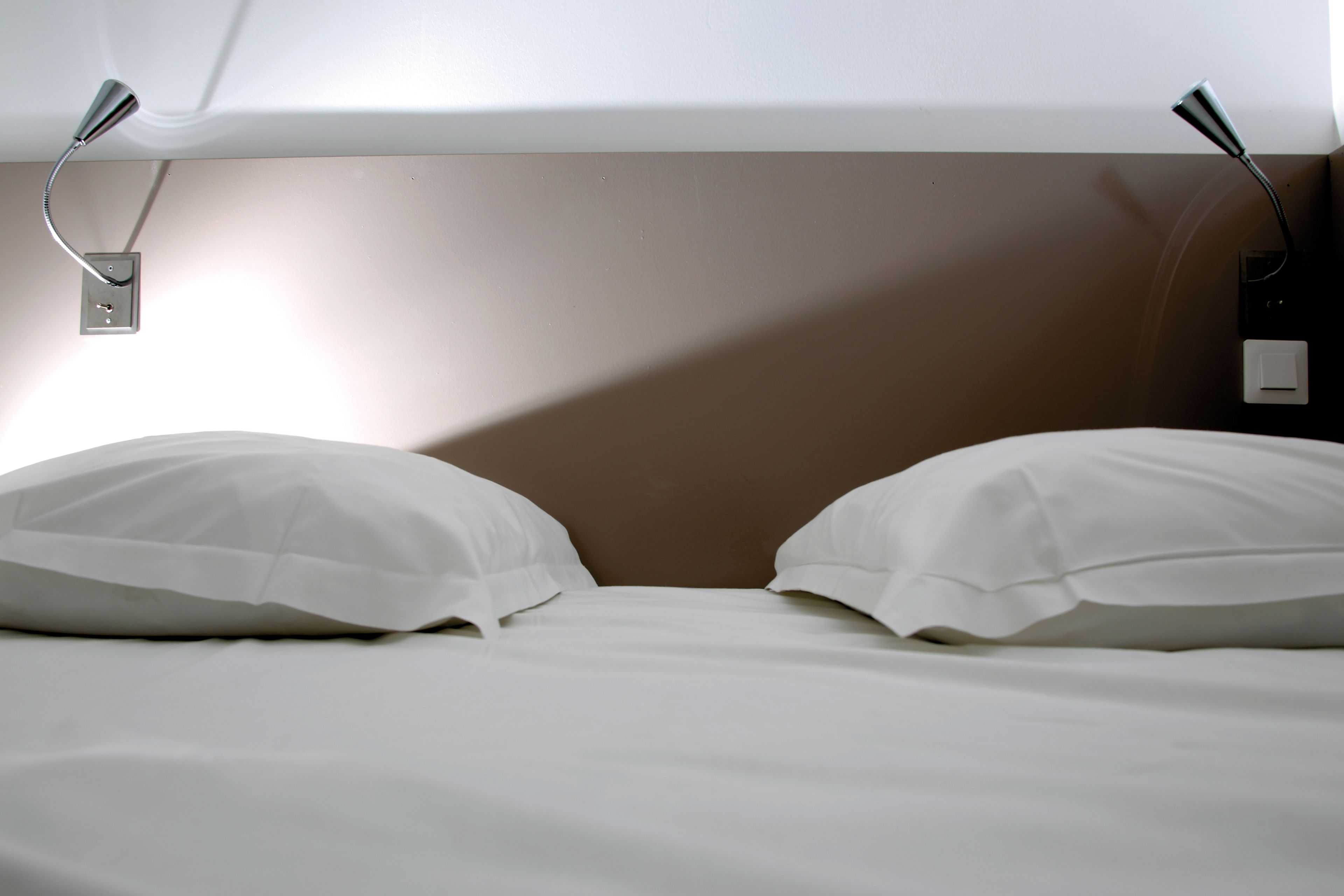 A bed with white bedding and two white pillows, a brown headboard, and a wall-mounted light fixture.