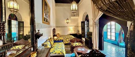 A traditional Moroccan room with a bed, a wooden table, a mirror, and a view of the outside.