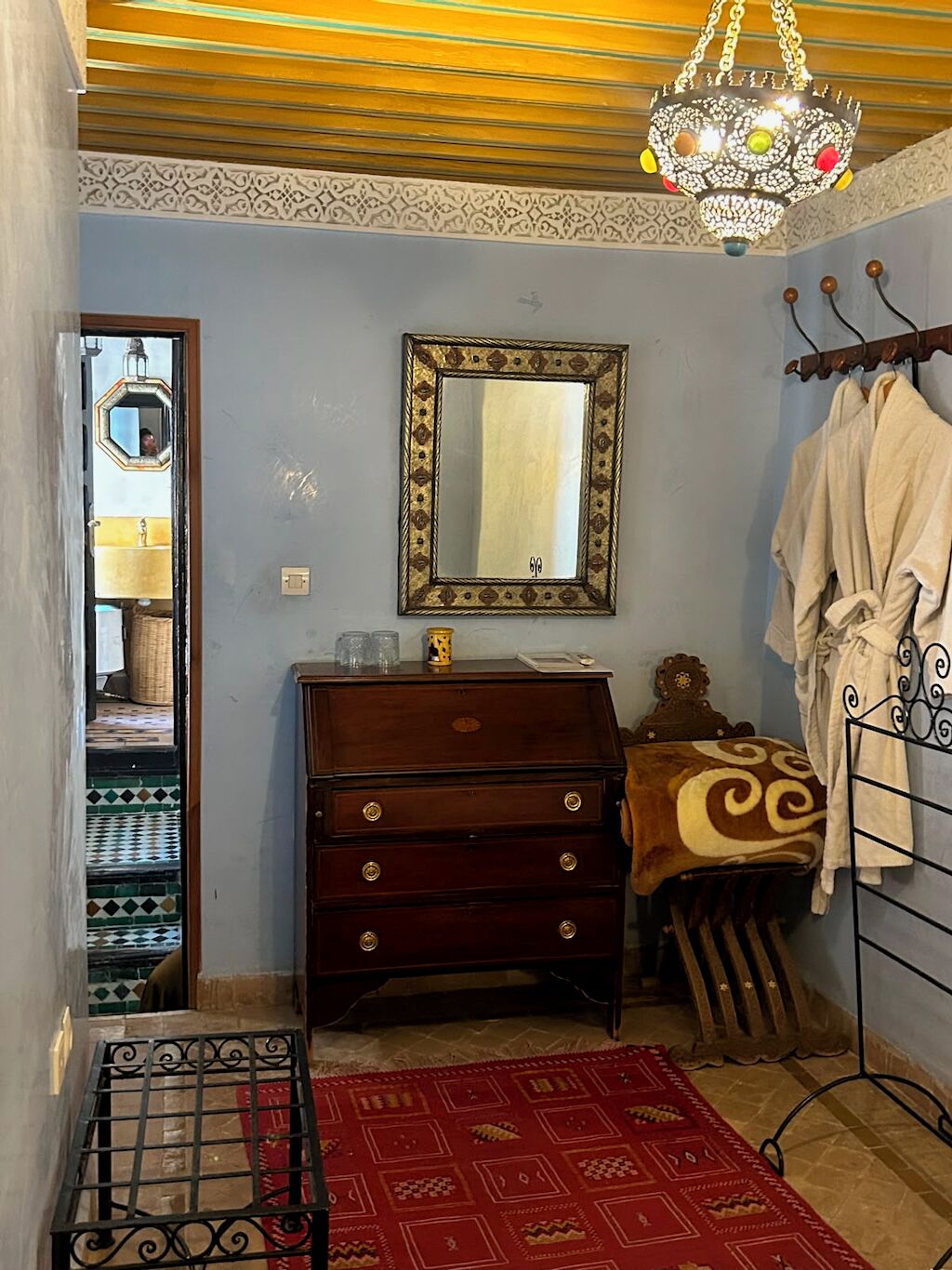 A room with a wooden dresser, a mirror with a decorative frame, a red patterned rug, and a metal rack with towels.
