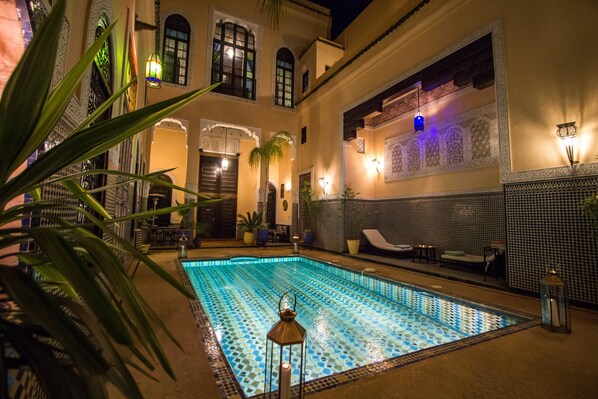A swimming pool with a tiled pattern, surrounded by a courtyard with palm trees and a seating area.