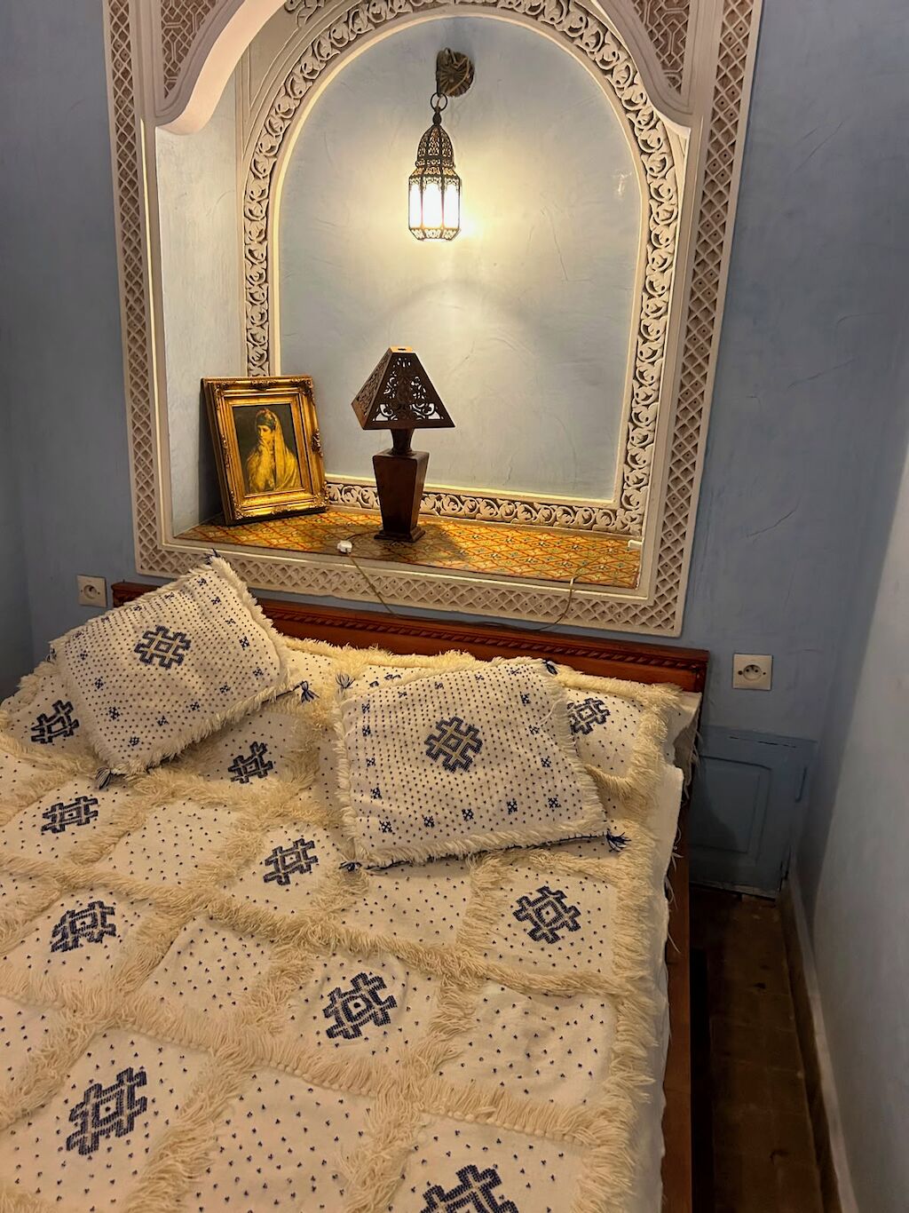 Ein Schlafzimmer mit einem Bett, Kissen, einem Nachttisch, einer Lampe und einem Spiegel mit dekorativem Rahmen.