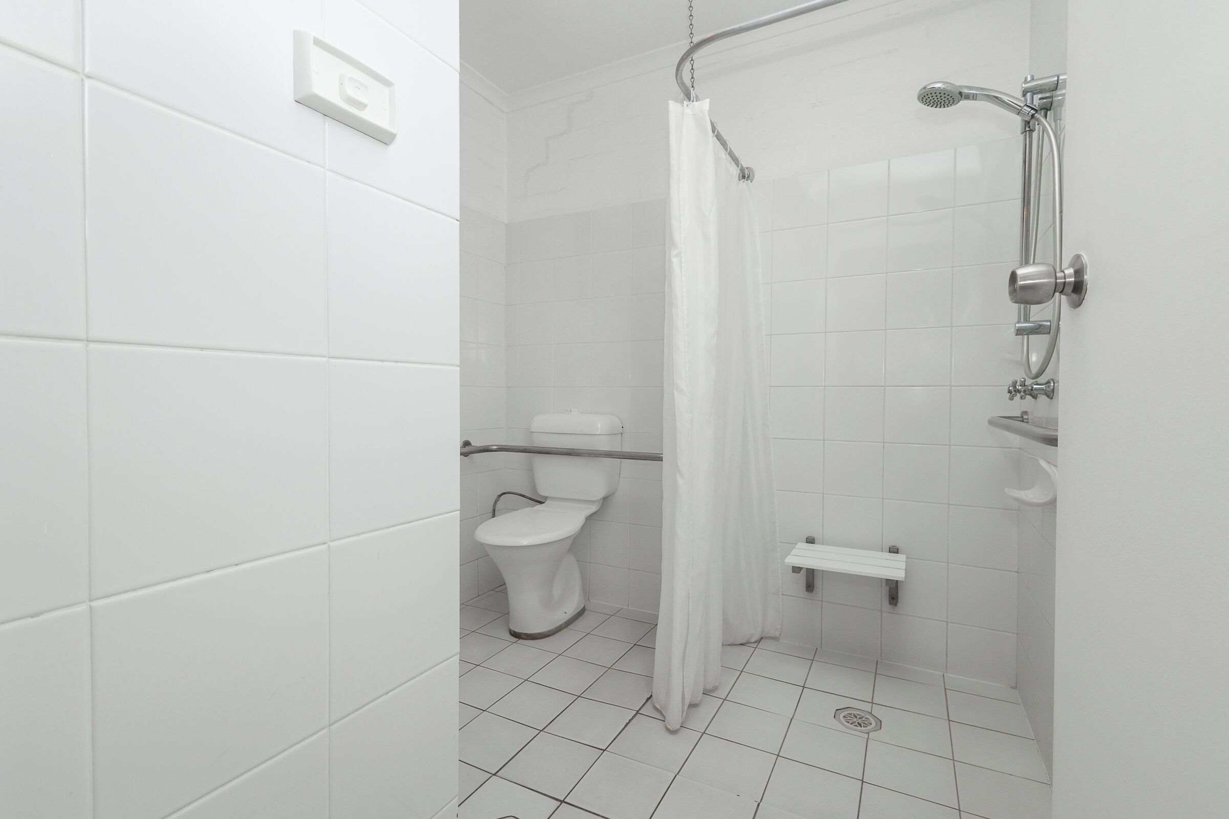 Un baño con azulejos blancos, un inodoro, una ducha con cortina y un estante pequeño.