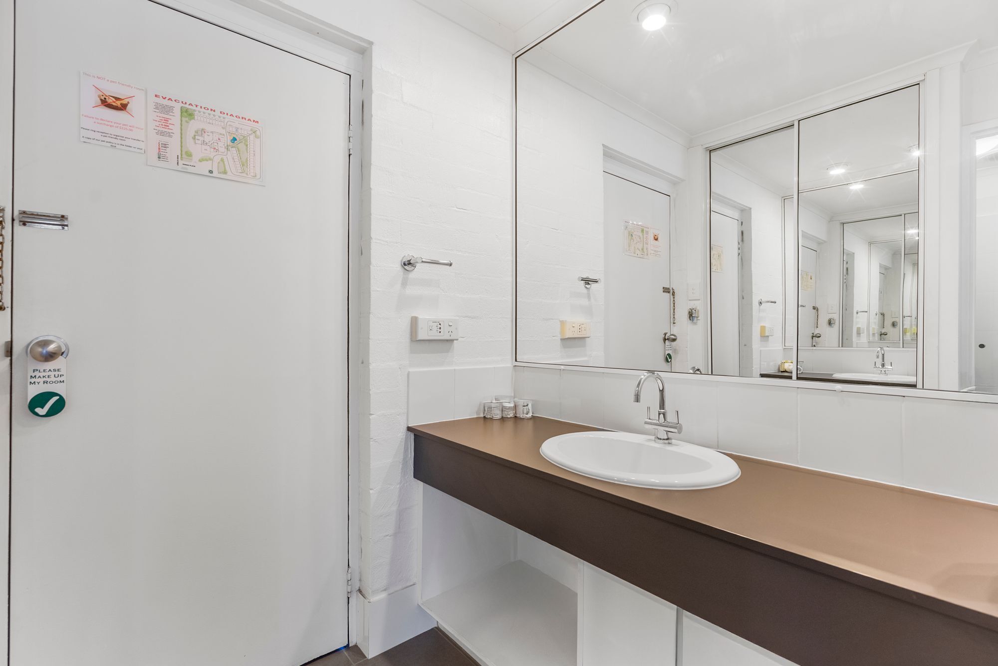 A modern bathroom with a white door, a "Please Make Up My Room" sign, a large mirror, and a sink with a brown countertop.