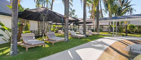 Un área junto a la piscina con sillones de descanso y sombrillas, rodeada de palmeras y un edificio con techo de metal.