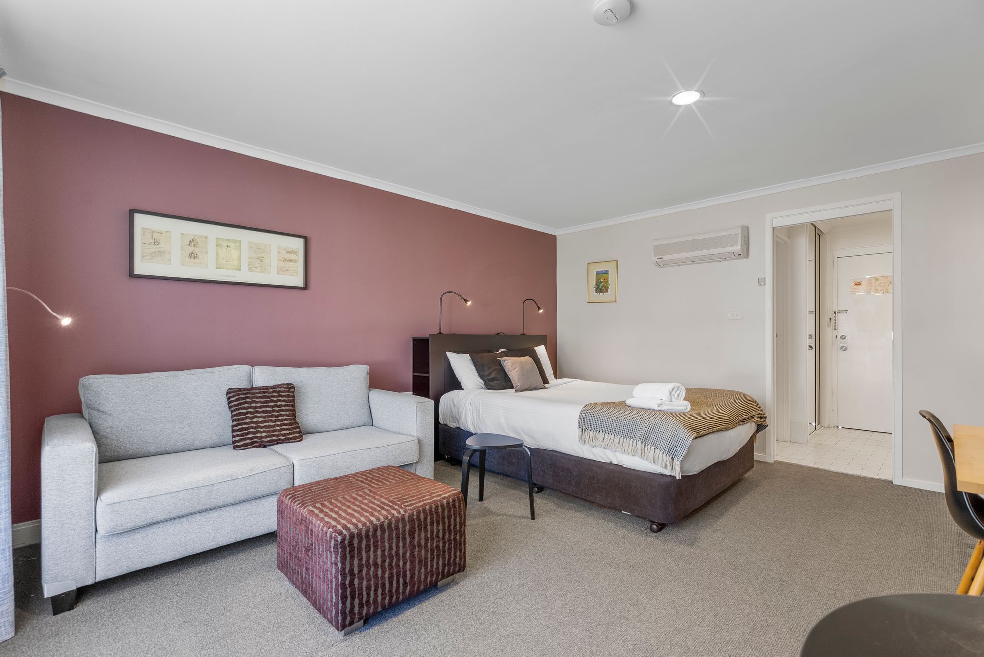 Une chambre d’hôtel avec un canapé gris, un lit recouvert d’une courtepointe à motifs, un petit pouf et un certificat encadré accroché au mur.