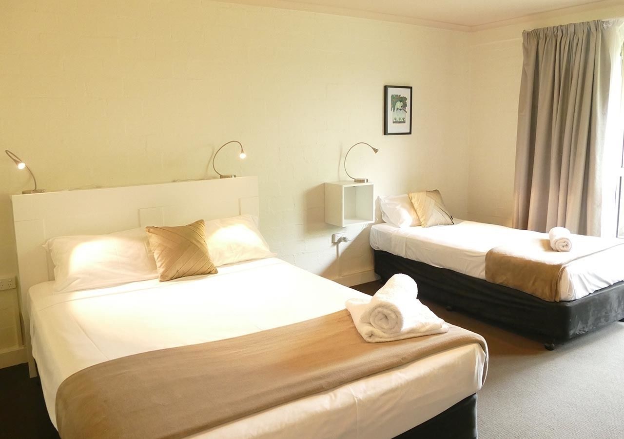 A hotel room with two beds, each with white linens and a beige throw blanket. There are bedside lamps and a framed picture on the wall.