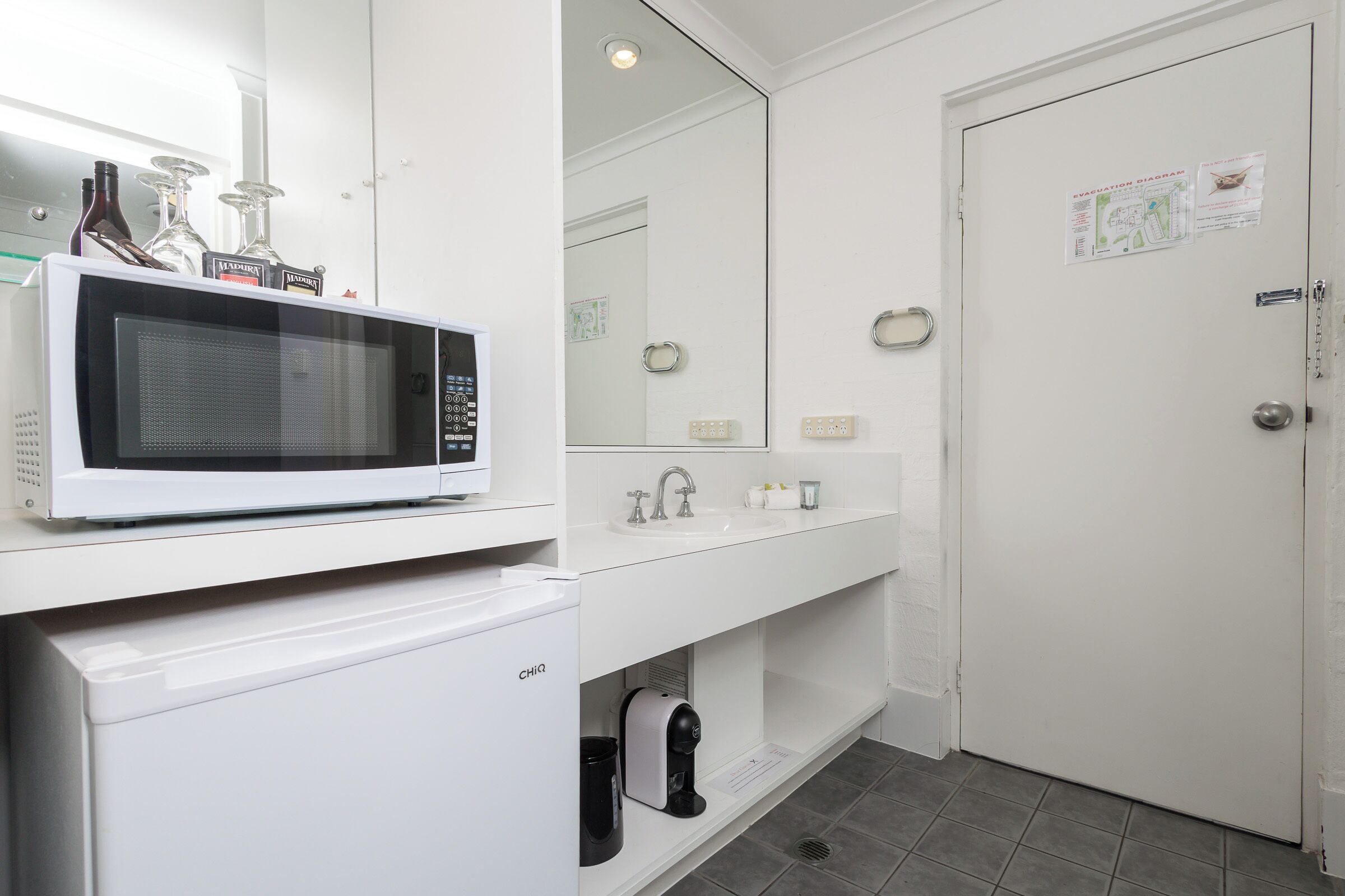A modern bathroom with a white sink, a mirror, a microwave, and a coffee machine.