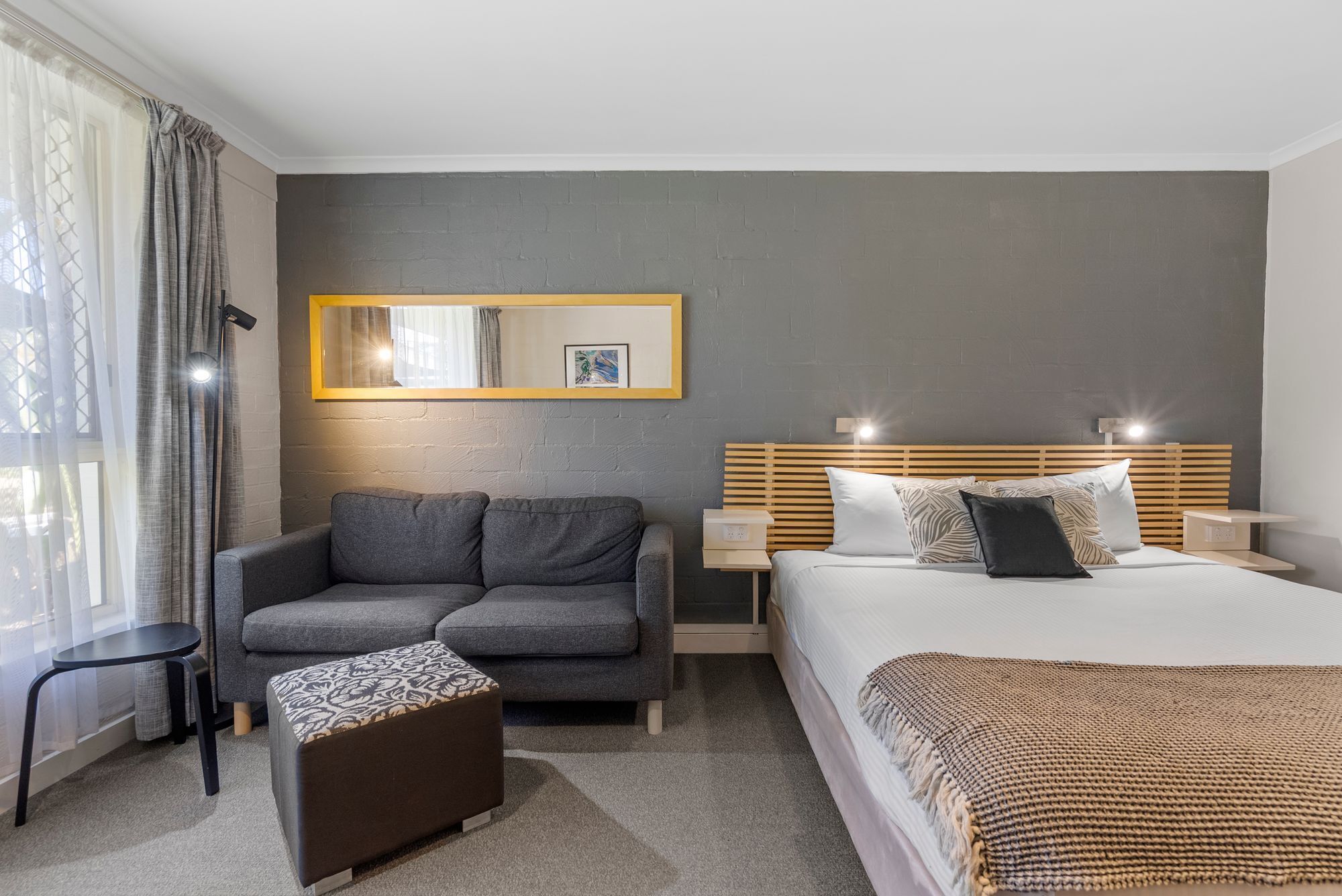 A modern hotel room with a grey sofa, a small ottoman, a bed with a wooden headboard, and a nightstand with a lamp.