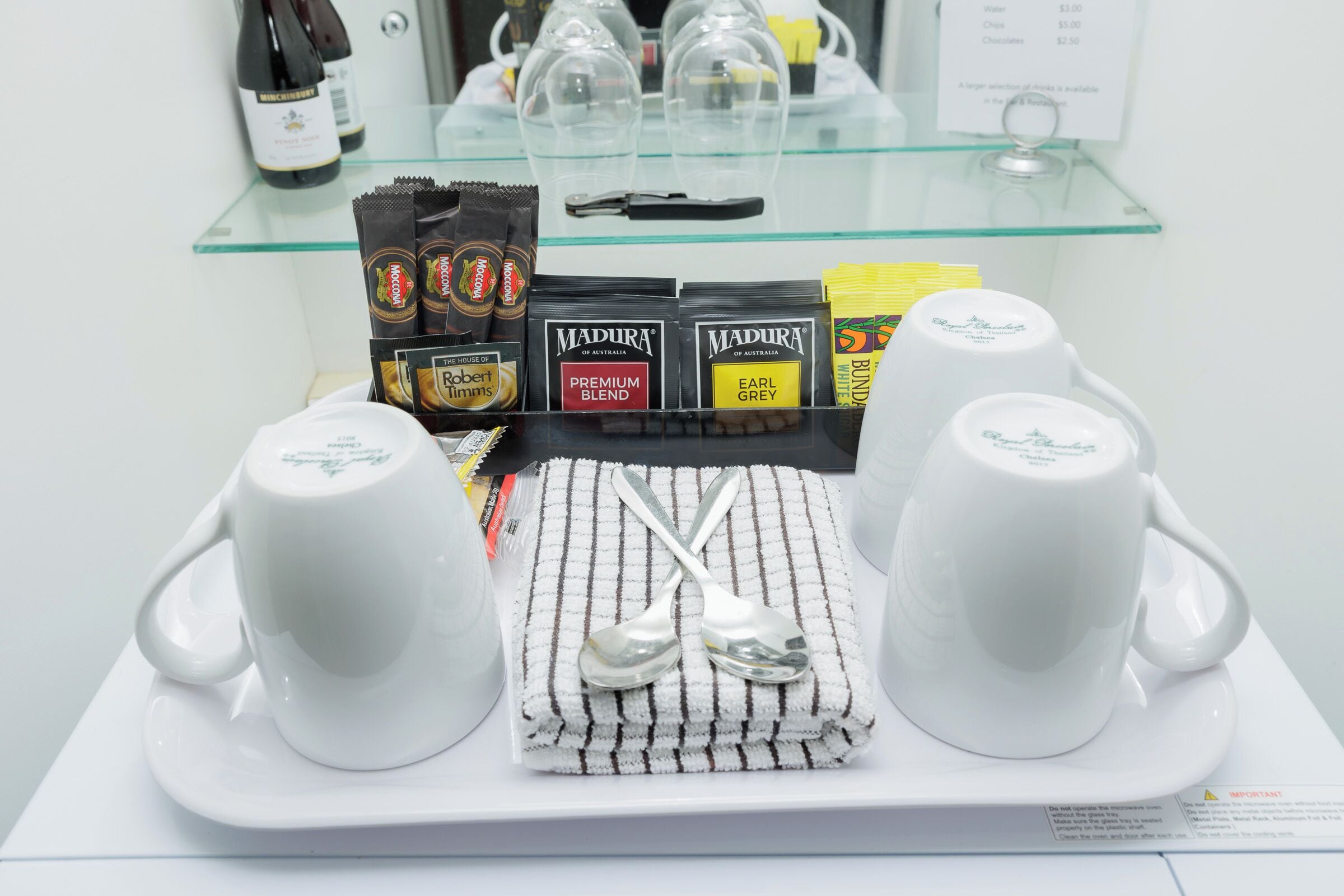 Una bandeja con dos tazas blancas y dos cucharas sobre una mesa blanca, con barras de chocolate y una botella de vino al fondo.
